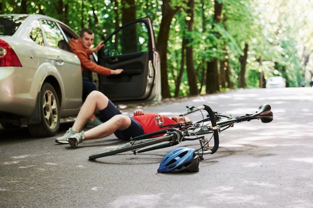 Conductor abriendo la puerta. Víctima en el asfalto. Bicicleta y accidente de coche de color plateado en la carretera en el bosque durante el día