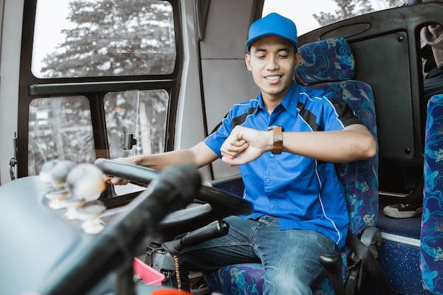 Un conductor de autobús masculino en uniforme azul mira su reloj mientras está sentado conduciendo en el autobús