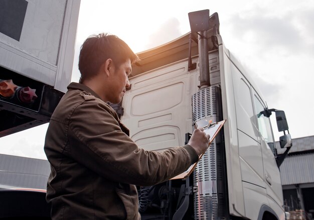 Conductor de camión asiático con portapapeles inspeccionando la lista de verificación de mantenimiento del vehículo de seguridad de semi camión moderno