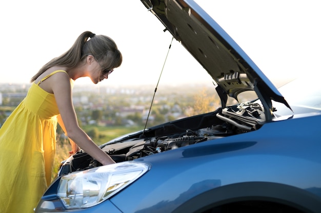 Foto conductor desconcertado de la mujer de pie cerca de su coche con el capó levantado inspeccionando el motor roto.