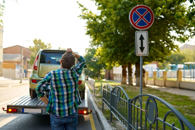 Conductor hombre que parece preocupado por tener una violación debido a un estacionamiento inadecuado. Camión de remolque que transporta su coche