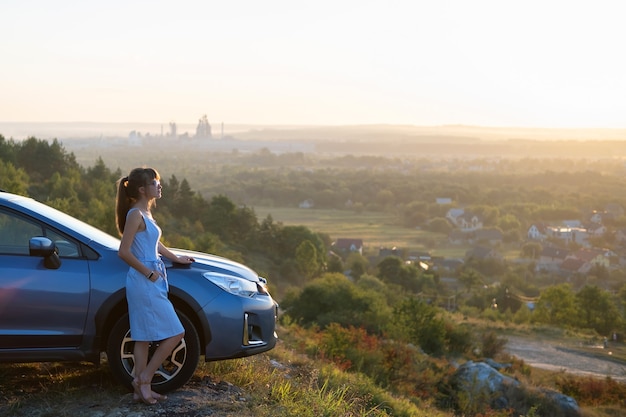 Foto conductor joven feliz en vestido azul disfrutando de una cálida noche de verano de pie junto a su coche. concepto de viajes y vacaciones.