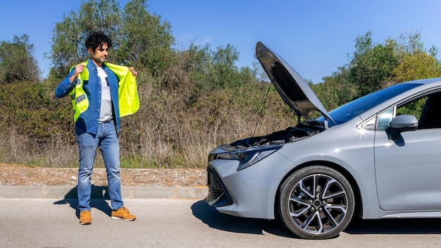 Conductor joven parado cerca de un auto roto con el capó levantado esperando ayuda