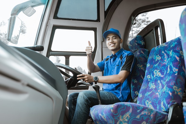 Un conductor masculino en uniforme azul sonríe a la cámara con un pulgar hacia arriba mientras está sentado en el autobús