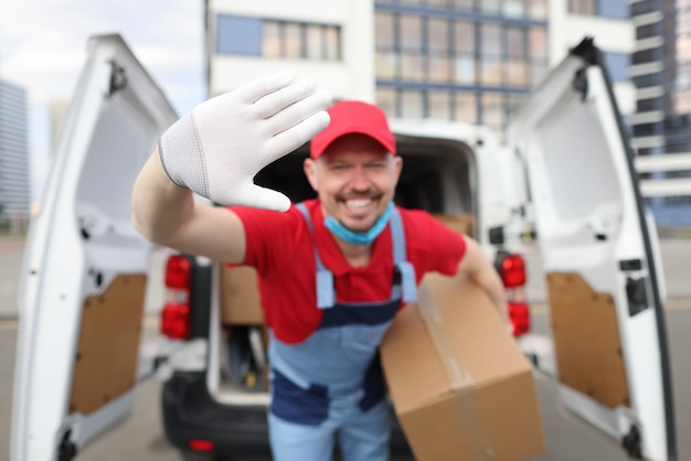 El conductor de mensajería en uniforme está sonriendo y saludando en la mano sosteniendo la caja de corton en la mano