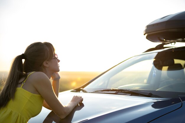 Conductor de mujer feliz en vestido de verano disfrutando de una cálida noche cerca de su coche. Concepto de viajes y vacaciones.