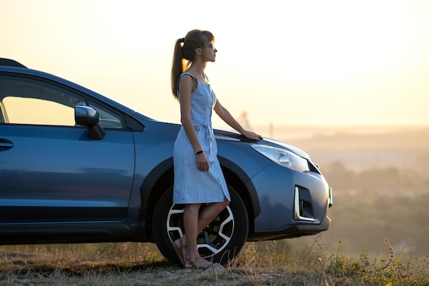 Conductor de mujer joven feliz en vestido azul apoyado en su coche disfrutando de un cálido día de verano. Concepto de viajes y vacaciones.