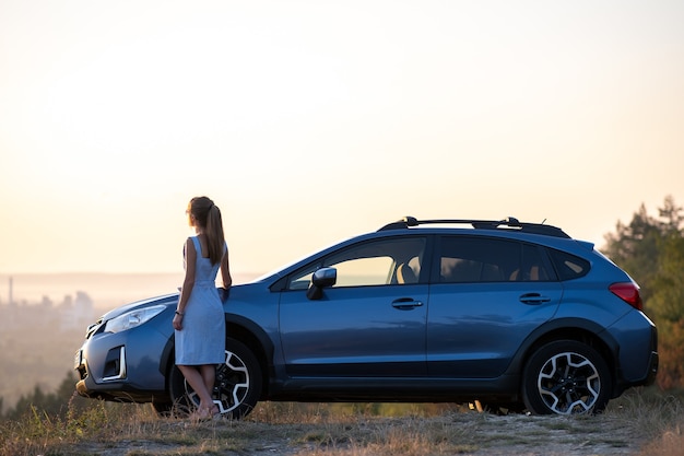 Conductor de mujer joven feliz en vestido azul disfrutando de una cálida noche de verano de pie junto a su coche. Concepto de viaje y vacaciones.