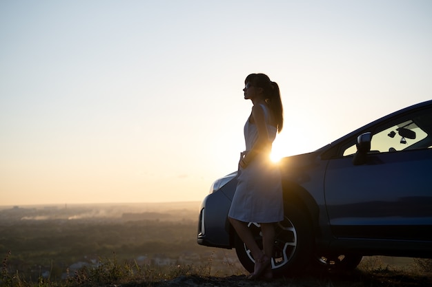 Foto conductor de mujer joven feliz en vestido azul disfrutando de una cálida noche de verano de pie junto a su coche. concepto de viaje y vacaciones.