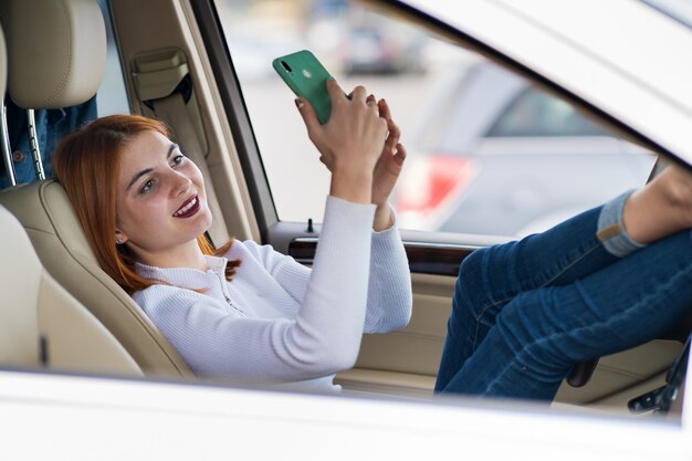 Conductor de mujer joven pelirroja tomando selfies con su teléfono móvil sentado al volante del coche en hora punta atasco de tráfico.