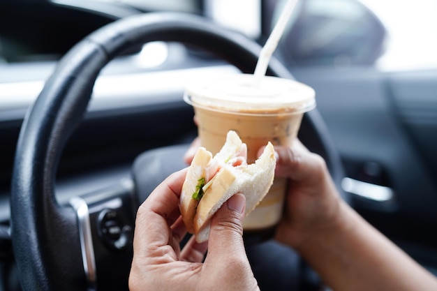 La conductora asiática sostiene una taza de café helado y pan de sándwich para comer y beber en un auto peligroso