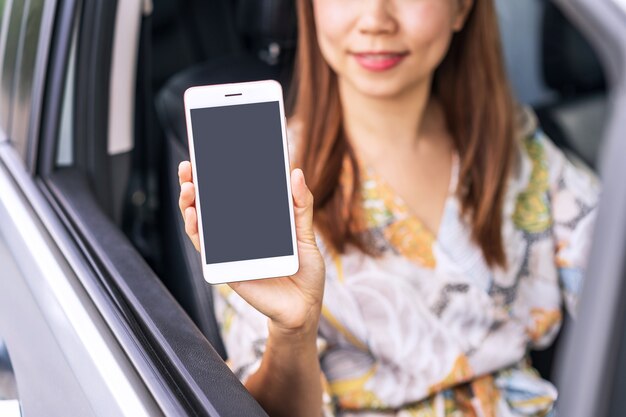 Conductora joven sentada en un coche y mostrando un teléfono inteligente de pantalla vacía