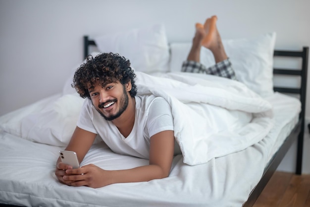 Conectado. Sonriente apuesto hombre indio de pelo rizado con un teléfono inteligente en las manos