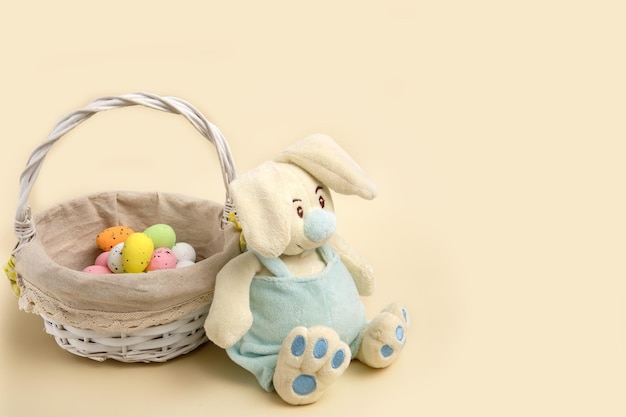 El conejito de pascua se sienta junto a una canasta con huevos coloridos sobre un fondo beige con espacio para copiar