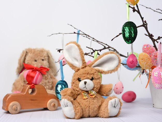 conejo de peluche sentado en un fondo blanco y huevos de Pascua decorativos de colores colgados en una rama