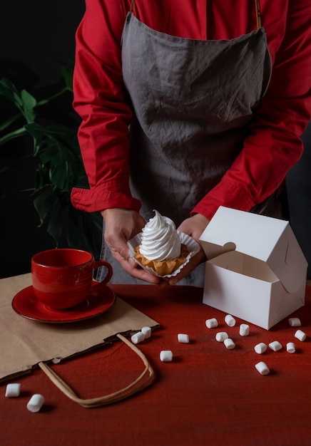 Confeiteiro segurando uma caixa de papel branco com bolo branco perto da mesa vermelha