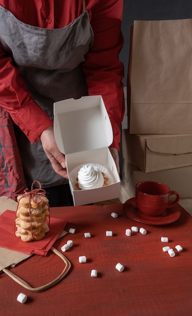 Confeiteiro segurando uma caixa de papel branco com bolo branco perto da mesa vermelha.