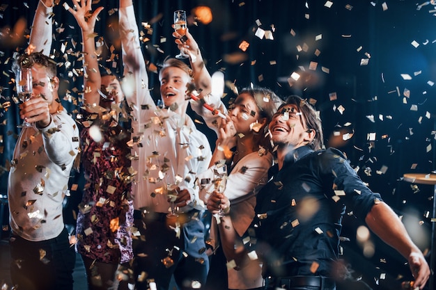 El confeti está en el aire. Grupo de amigos alegres celebrando el año nuevo en el interior con bebidas en las manos.