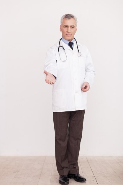 ¡Confía en mí! Longitud total de médico senior de cabello gris en uniforme blanco mirando a la cámara y estirando la mano mientras está de pie aislado en blanco