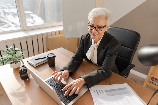 Confiada y elegante mujer mayor europea de mediana edad que usa una computadora portátil en el lugar de trabajo