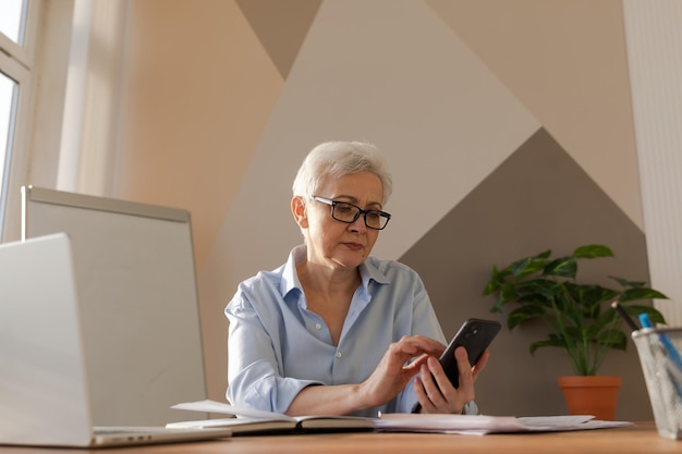 Confiada y elegante mujer mayor europea de mediana edad que usa un teléfono inteligente en el lugar de trabajo con estilo matu mayor