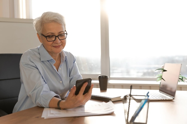 Confiada y elegante mujer mayor europea de mediana edad que usa un teléfono inteligente en el lugar de trabajo con estilo matu mayor