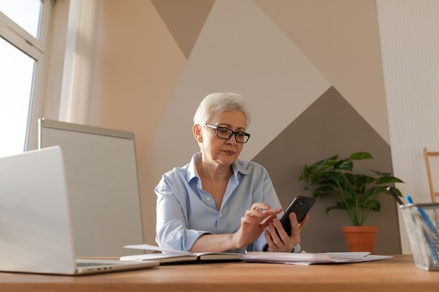 Confiada y elegante mujer mayor europea de mediana edad que usa un teléfono inteligente en el lugar de trabajo con estilo matu mayor