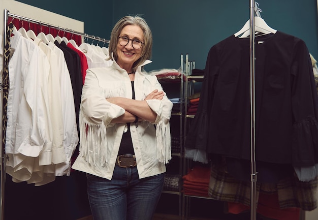 Confiada y exitosa mujer caucásica madura costurera modista sastre posando en su propio taller de diseño de moda Diseñadora de ropa entre perchas con prendas de vestir en taller de sastrería