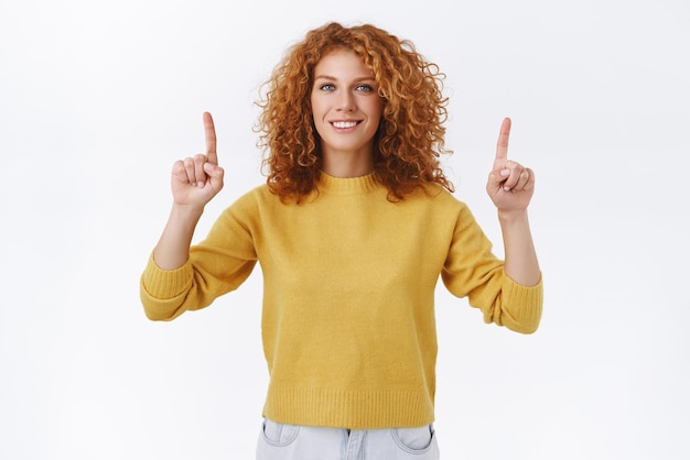 Confiada y hermosa mujer pelirroja sonriente con cabello rizado, usa suéter amarillo, levanta los brazos y señala hacia arriba, da la dirección donde colgar la pintura te invita a visitar el evento o la fiesta que muestra la promoción