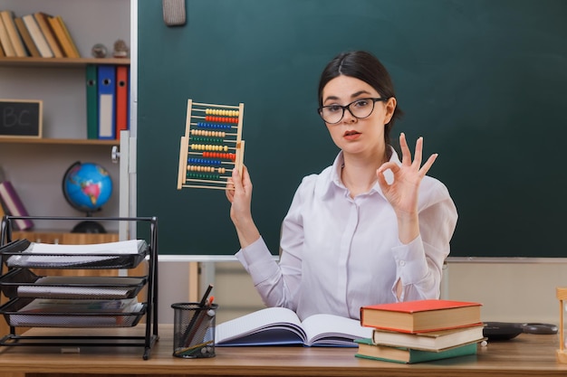 confiada mostrando un buen gesto joven maestra con gafas sosteniendo abacus sentado en el escritorio con herramientas escolares en el aula