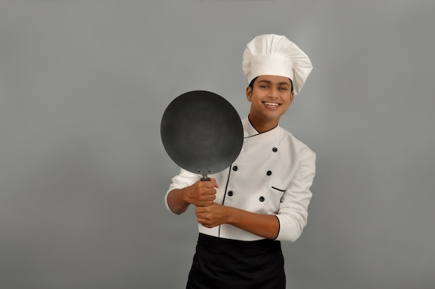 Confiado chef indio sonriente sonriendo detrás de la sartén sobre fondo gris