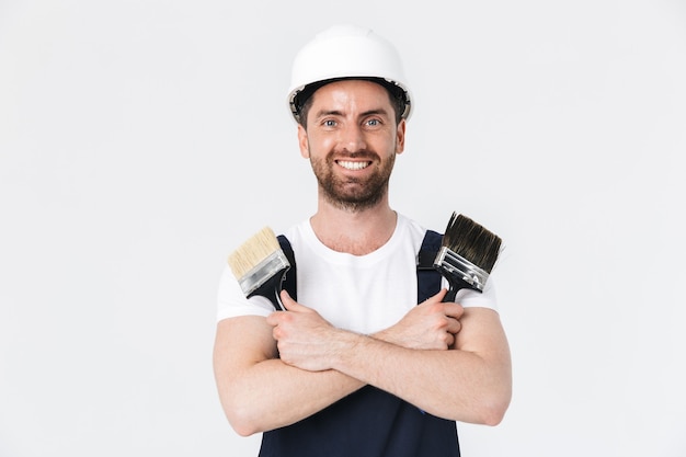 Confiado constructor barbudo hombre vestido con monos y casco que se encuentran aisladas sobre la pared blanca, mostrando pinceles