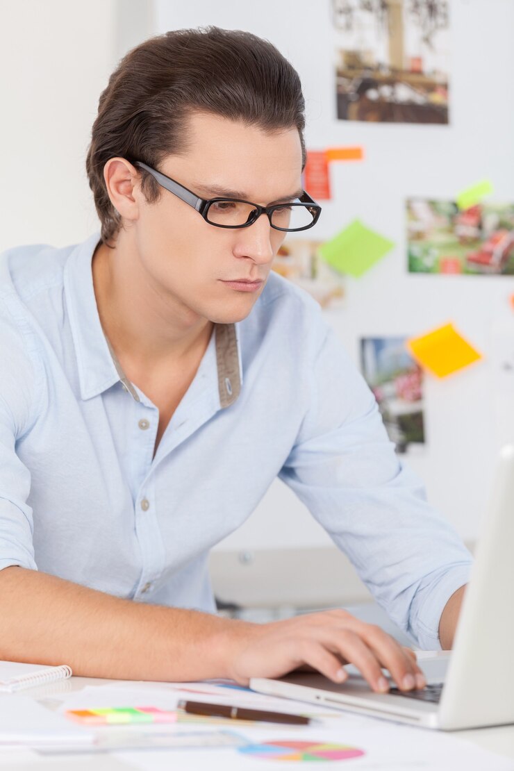 Foto confiado y creativo. hombre joven pensativo en camisa azul trabajando en equipo mientras está sentado en su lugar de trabajo