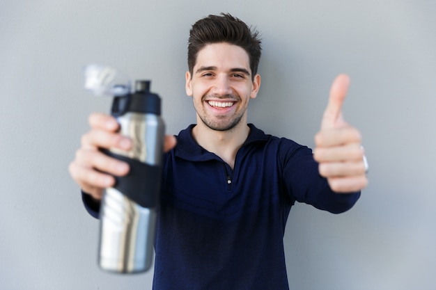 Foto confiado deportista sonriente sosteniendo una botella de agua mientras está de pie aislado sobre fondo gris