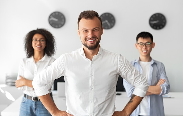 Confiado empresario barbudo con las manos en la cintura sonriendo para la cámara mientras es líder de un equipo multirracial durante el trabajo en la oficina moderna