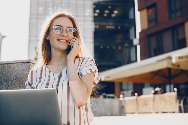 Confiado empresario caucásico con pelo rojo y pecas está hablando con alguien por teléfono mientras usa una computadora afuera