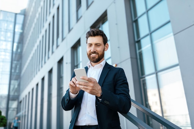 Confiado empresario milenario caucásico con barba en traje escribiendo en smartphone chateando y navegando en internet