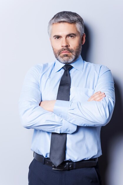 Confiado y exitoso. Hombre maduro confiado en camisa y corbata mirando a la cámara y manteniendo los brazos cruzados mientras está de pie contra el fondo gris