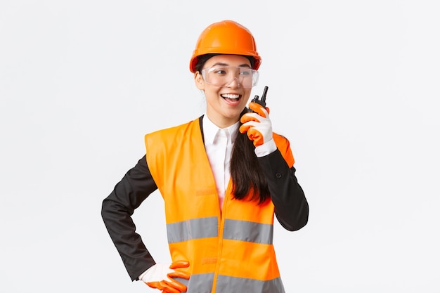 Confiado ingeniero asiático complacido en casco de seguridad y uniforme hablando con el arquitecto jefe usando walkie-talkie. Equipo de contacto técnico de construcción satisfecho mediante teléfono de radio