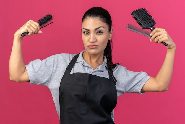 Foto confiado joven barbero mujer caucásica vistiendo uniforme mirando al frente sosteniendo peines haciendo gesto fuerte aislado en la pared rosa