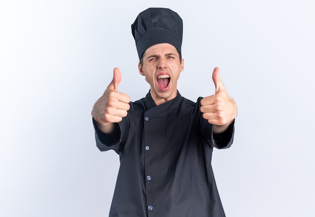 Confiado joven cocinero de sexo masculino rubio en uniforme de chef y gorra mirando a la cámara mostrando los pulgares para arriba gritando aislado en la pared blanca