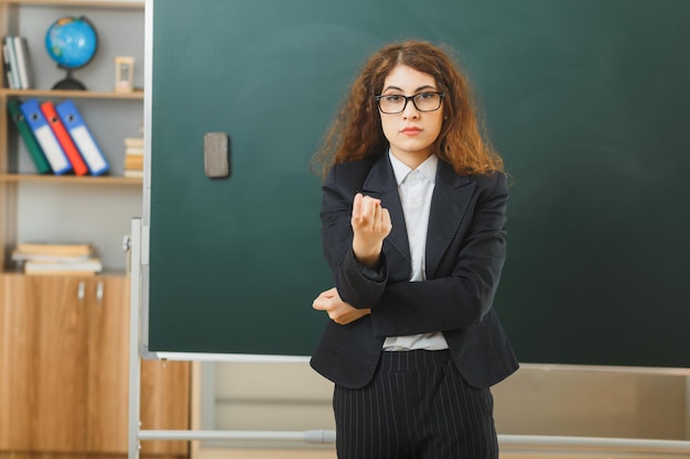 Confiado mostrando gesto joven maestra de pie frente a la pizarra en el aula