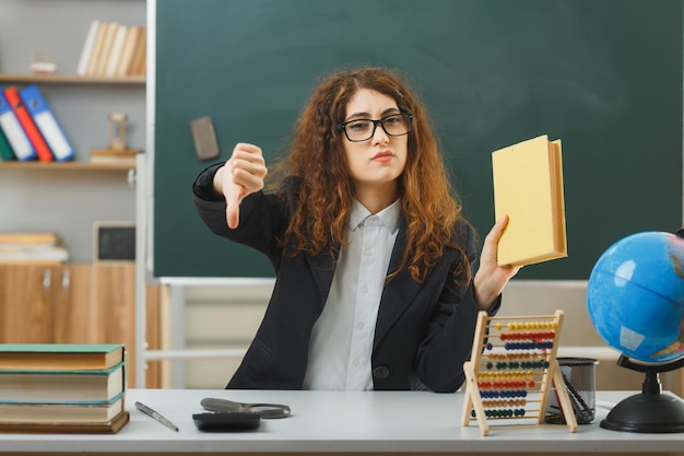 confiado mostrando pulgares hacia abajo joven maestra con gafas sosteniendo un libro sentado en el escritorio con herramientas escolares en el aula