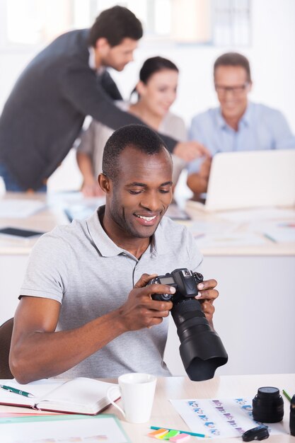 Confiado en su trabajo. Hombre africano joven confidente que sostiene la cámara y sonriendo mientras tres personas trabajan en segundo plano