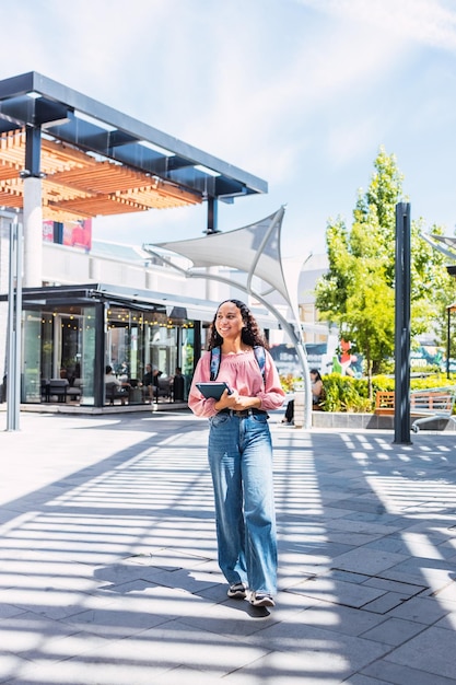 Confiança. Estudante universitária latina sorridente andando e segurando seus livros fora do shopping