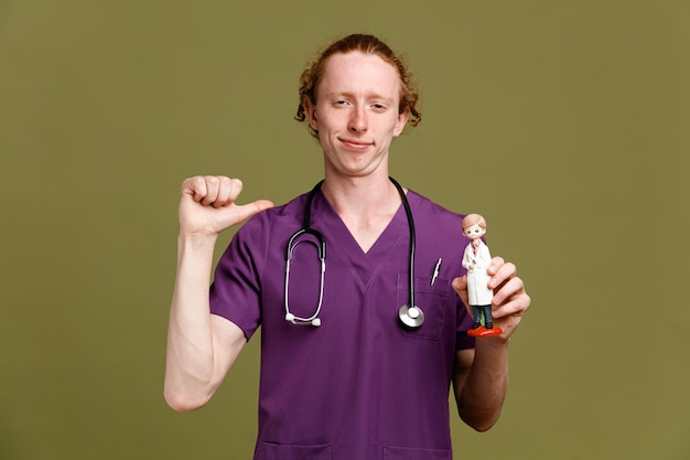 Confiante aponta para si mesmo jovem médico masculino vestindo uniforme com estetoscópio segurando brinquedo isolado em fundo verde