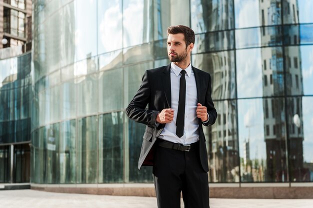 Confiante e bem-sucedido. Bonito e jovem empresário ajustando o paletó e desviando o olhar enquanto caminhava ao ar livre com o prédio de escritórios ao fundo