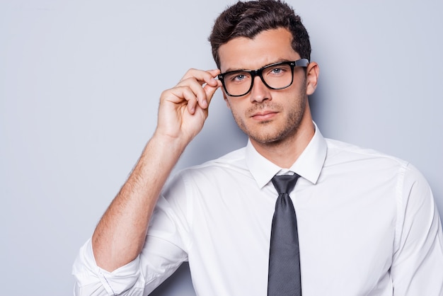 Confiante e bem-sucedido. Retrato de jovem bonito de camisa e gravata ajustando os óculos e olhando para a câmera em pé contra um fundo cinza