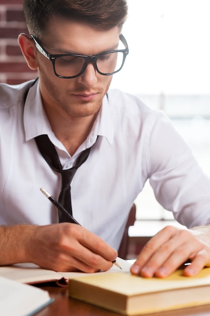 Confiante e criativo. Jovem bonito de camisa e gravata escrevendo algo no bloco de notas enquanto está sentado em seu local de trabalho