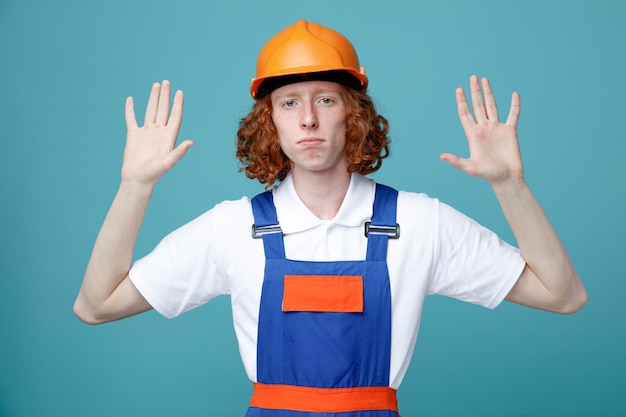 Confiante espalhando as mãos jovem construtor de uniforme isolado em fundo azul